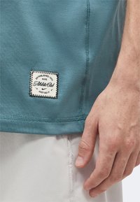 Κοντινή λήψη χεριού δίπλα στο βεραμάν τελείωμα μπλουζάκι Nike Athletic Club με ραμμένη λευκή ετικέτα και ανοιχτόχρωμο σορτς.