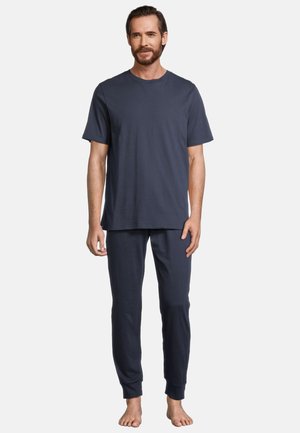 Homme debout pieds nus portant une chemise bleu foncé à manches courtes et un pantalon de jogging assorti sur fond blanc.
