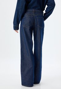 Jeans de mezclilla de pierna ancha en azul oscuro, con cintura alta, dos bolsillos traseros y costura visible a lo largo de las costuras. Textura suave.