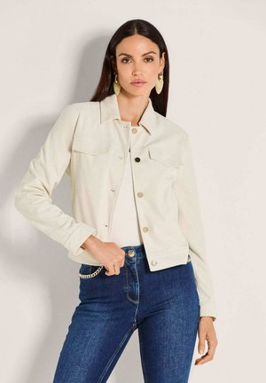 Frau mit langen dunklen Haaren, die eine cremefarbene, geknöpfte Jacke, ein weißes Oberteil, blaue Jeans mit Kettendetail und große ovale Ohrringe vor hellem Hintergrund trägt.