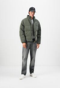 Homme portant une veste matelassée verte, un T-shirt gris, un jean noir délavé, des baskets blanches et une casquette de baseball noire, debout contre un fond blanc.