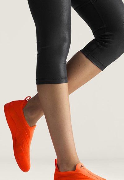 Ben iført sorte korte leggings og lyse orange træningssko med Adidas-logoet og teksten "Stella McCartney".