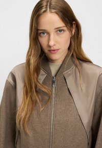 Ruskea pommittaja-takki, jossa on villainen vartalo, kiiltävät vaaleanruskeat hiha-akcentit, ribbauskaulus ja metallivetoketju. Yksinkertainen, moderni muotoilu, jossa on selkeät linjat.