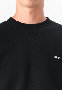 Črno pulover s krožnim ovratnikom, rebrastimi manšetami in majhnim belim logotipom "OBEY" spredaj. Gladka, mehka tekstura tkanine.