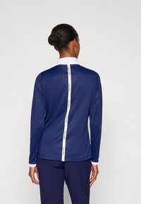 Námořnicky modrá polokošile s dlouhým rukávem, bílým límečkem a vertikálním pruhovým logem na zádech. Hladká látka a přiléhavý střih.