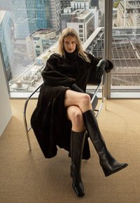 Naine, kes kannab musta karvkattega mantlit, musti kindaid ja põlveni ulatuvaid musta nahast saapaid, istub jalad ristatud moodsa linnavaatega kontoris.