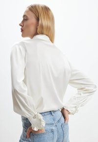 White satin blouse with a high collar, gathered sleeves, and decorative button accents, paired with high-waisted blue denim shorts.