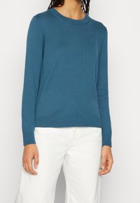 Blauer Strickpullover mit langen Ärmeln und gerippten Abschlüssen. Verfügt über einen klassischen Rundhalsausschnitt und ein dezentes Logo auf der Brust. Kombiniert mit weißen Hosen.