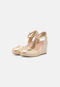 Pár zlatých metalických espadril na wedges s kotníkovými pásky a texturovanými tkanými podpatky na bílém pozadí.