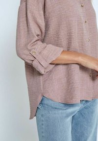 Light pink button-up shirt in textured fabric with rolled sleeves and side slits, paired with blue jeans.