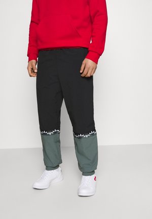 Persona vistiendo pantalones bloqueados en negro y gris con ribete blanco de logo, zapatillas blancas y una sudadera con capucha roja sobre un fondo liso.