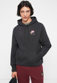 Jovem com cabelo encaracolado escuro, vestindo um hooded da Nike Air preto e calças de moletom bordô, segurando um boné preto com a mão esquerda.