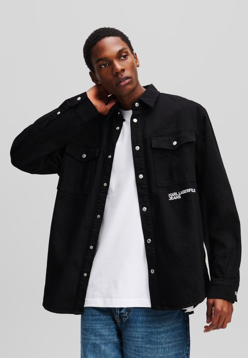 Chemise noire surdimensionnée en coton, dotée de deux poches poitrine et de boutons-pression argentés. Logo blanc imprimé sur le côté gauche.