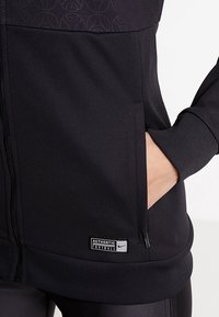Gros plan sur une main dans la poche latérale d'une veste noire zippée avec une étiquette "Authentic Football" près de l'ourlet.