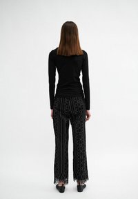 Schwarzes Langarmoberteil kombiniert mit schwarzen Hosen, die ein vertikales Streifenmuster, kreisförmige Designs und eine Spitzenborte am Saum aufweisen.