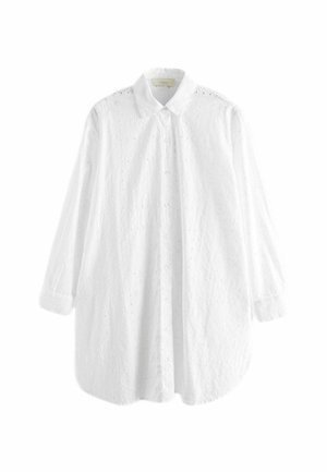 Femme portant une robe-chemise blanche à manches longues brodée d'œillets près d'une piscine, avec manches retroussées et bracelets au poignet gauche.