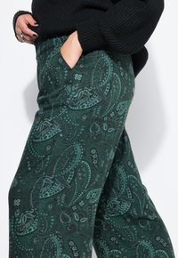Personne portant un pantalon à motifs paisley vert foncé avec un waistband élastique, un pull noir, les mains dans les poches, sur fond blanc.