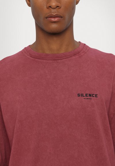 Rövid ujjú, bordó környakú póló, puha pamutból készült, elhalványult textúrával. A frontján fekete logó található, amely a "SILENCE" feliratot viseli.
