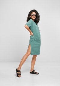 Muntgroene jurk met korte mouwen, zijsplitten en een zak. Gecombineerd met zwarte sandalen en ronde zonnebrillen. Gladde, zachte stoftextuur.