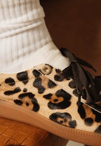 Sneaker with leopard print, black laces, white tongue, worn with white ribbed sock on a brown textured surface.