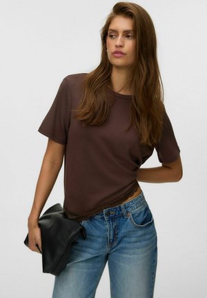 Vrouw met lang bruin haar draagt een bruin kortärmelig topje en blauwe spijkerbroek, en houdt een zwarte gevouwen clutch vast tegen een effen achtergrond.