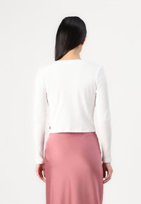 Haut blanc à manches longues côtelées avec un décolleté carré, conçu court, et un petit logo rouge à l'arrière, associé à une jupe fluide rose.