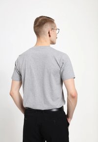 Šedé bavlněné tričko s krátkým rukávem, kulatým výstřihem a hladkou texturou. Nosené s černými kalhotami a černým pásem, pohled zezadu.