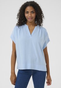 Blouse bleu clair à manches courtes avec un décolleté en V, coupe décontractée et un tissu légèrement texturé ; l'ourlet est droit et se termine à la taille.