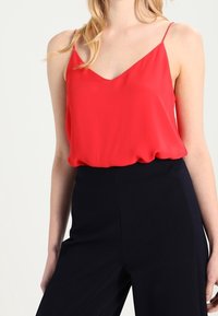 Donna che indossa una maglia rossa senza maniche a scollo a V infilata in pantaloni neri a vita alta, su uno sfondo chiaro e semplice.