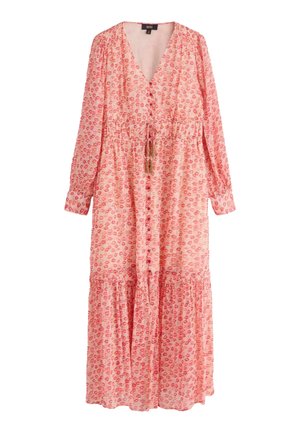 Kobieta idąca w zwiewnej czerwonej sukience we wzory z wysokim rozcięciem, do której dobrano jasne kowbojskie botki do kostki, na czerwonym tle.