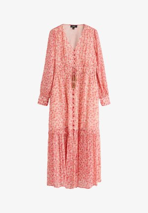 Nő sétál egy légies, mintás, piros ruhában, magas hasítékkal, világos bokacsizmával, piros háttér előtt.