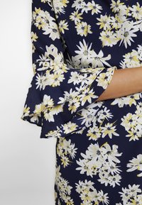 Blommig klänning med marinblå botten, prydd med vita och gula blommönster. Ärmarna är klockformade med lager av volanger och textil med struktur.