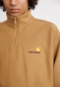 Tan quarter-zip sweatshirt made of soft fabric. Features a Carhartt logo embroidered in maroon and yellow on the chest. Simple design.