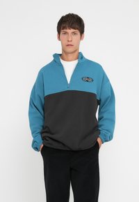 Quarter-zip genser i teal og svart fleece, med ribbestrikkede mansjetter, logo på brystet og avslappet passform. Myk og teksturert overflate.