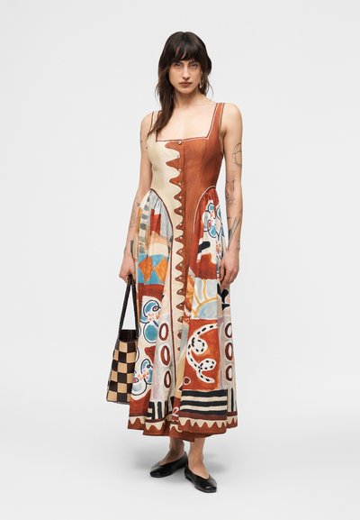 Γυναίκα με σκούρα μαλλιά που φοράει αμάνικο, midi φόρεμα με μοτίβο και κουμπιά, κρατώντας μια καφέ και μπεζ τσάντα με καρό σχέδιο.