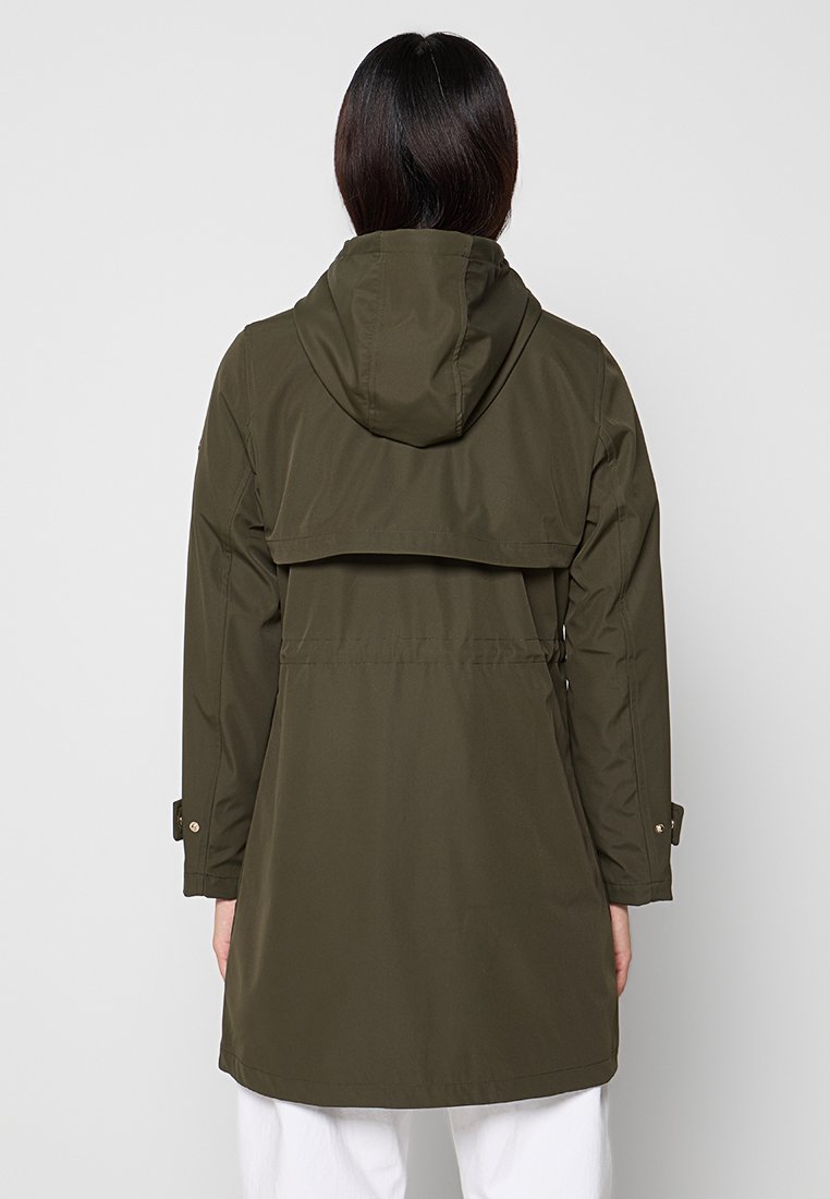 Kobieta z ciemnymi włosami, ubrana w oliwkową kurtkę przeciwdeszczową z kapturem, pokazana od tyłu, w białych spodniach na jednolitym tle.