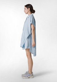 Light blue oversized shirt-dress with short sleeves, featuring a textured fabric and side slits, worn over a light blue layered garment.