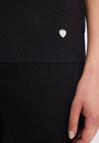 Zbliżenie ręki obok czarnego dzianinowego materiału z małym srebrnym charms'em w kształcie serca przymocowanym w pobliżu brzegu.