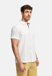 Camisa blanca de manga corta, con tela texturizada, cuello tradicional y detalle a rayas en contraste en la tapeta.
