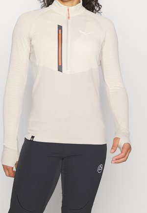 Persoon draagt een beige jack met kwartritssluiting en een oranje rits, en een zwarte broek met een wit logo op het bovenbeen, tegen een witte achtergrond.