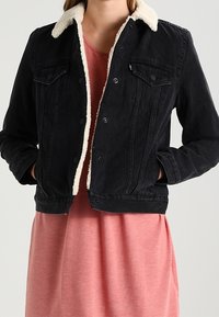 Chaqueta de mezclilla negra con cuello de sherpa color crema, cierre frontal con botones y dos bolsillos en el pecho, usada sobre un vestido rosa.