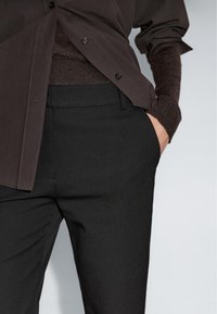 Persoon gekleed in zwarte taillepanten, een donkerbruin gebreid trui, en een donkerbruin knoopshirt met één hand in de broekzak.