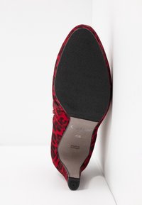 Chaussure à talon haut en velours rouge avec semelle noire, bout pointu et motif léopard, taille 4½, fabriquée au Portugal, présentée debout contre un coin blanc.