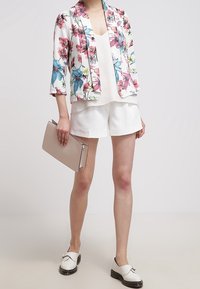 Marynarka w białym kolorze z nadrukiem kwiatowym w odcieniach niebieskiego i różowego, zestawiona z jasnym topem i białymi szortami. Do tego beżowa kopertówka jako dodatek.