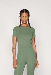 Olivově zelený sportovní top, krátké rukávy, těsný střih, hladká textura, viditelné švy. V kombinaci s odpovídajícími olivově zelenými legínami.