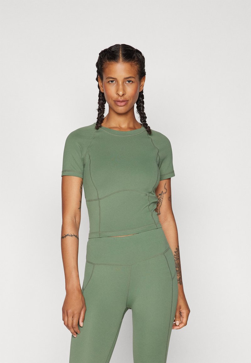 Olivově zelený sportovní top, krátké rukávy, těsný střih, hladká textura, viditelné švy. V kombinaci s odpovídajícími olivově zelenými legínami.