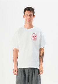 T-shirt en coton blanc à manches courtes et col rond. Présente un motif graphique rouge sur la poitrine. Associé avec un pantalon gris.