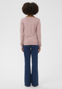 Hellrosa langärmliger Pullover mit rundem Ausschnitt und geripptem Saum, kombiniert mit ausgestellten blauen Denim-Jeans. Glatte Textur, figurbetontes Design.