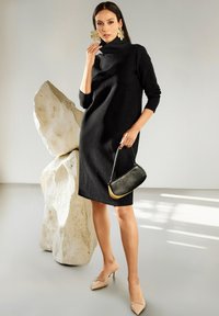 Frau in schwarzem Rollkragenkleid und nuden Absatzschuhen hält eine kleine schwarze Handtasche und steht neben einer großen weißen Steinskulptur in einem minimalistischen Ambiente.