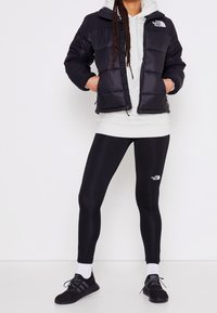 Chaqueta puffer negra con capucha, cierre de cremallera, combinada con una sudadera gris, leggings negros y zapatillas deportivas negras con detalles en blanco.